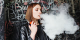 Young woman in a black leather jacket vaping, blowing a cloud of smoke. She stands against a graffiti-covered wall, conveying a rebellious mood.