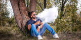 Man with sunglasses vapes while sitting against a tree, surrounded by greenery. He wears a dark shirt and jeans, exuding a relaxed, tranquil vibe.