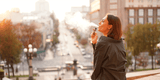 A woman stands on a city street, exhaling vapor from an e-cigarette. It's a sunny day, and her relaxed posture conveys a sense of calm and contentment.